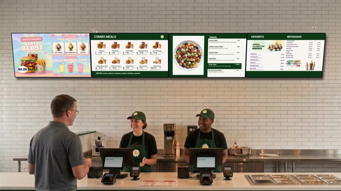 A restaurant counter with a wall of four digital menu boards above it: combos, salads and pizza, a featured burger, and drinks and shakes. A customer is ordering from two smiling staff.